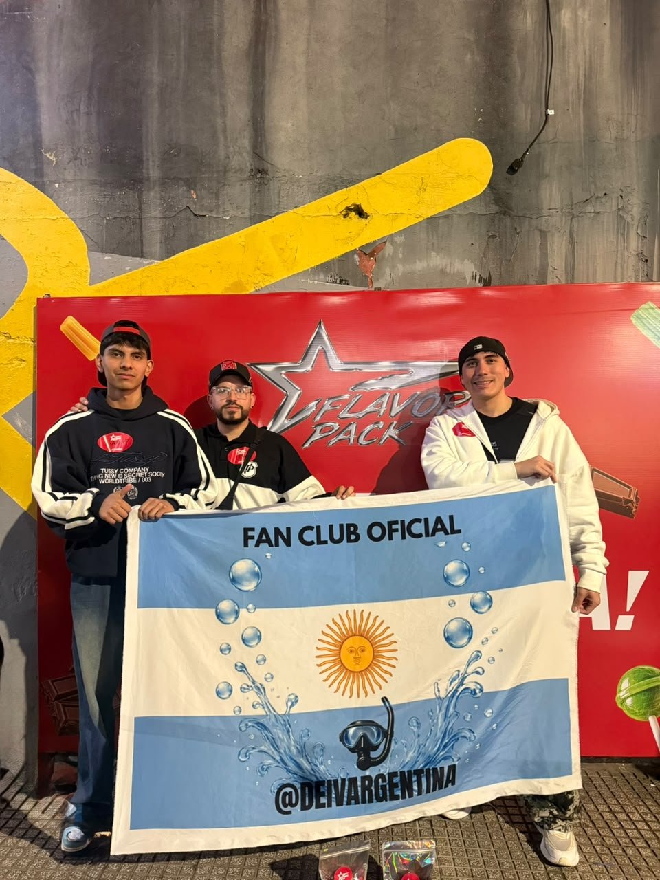Tres integrantes del fan club con la bandera Fan Club Oficial frente al cartel rojo del Flavorz Pack